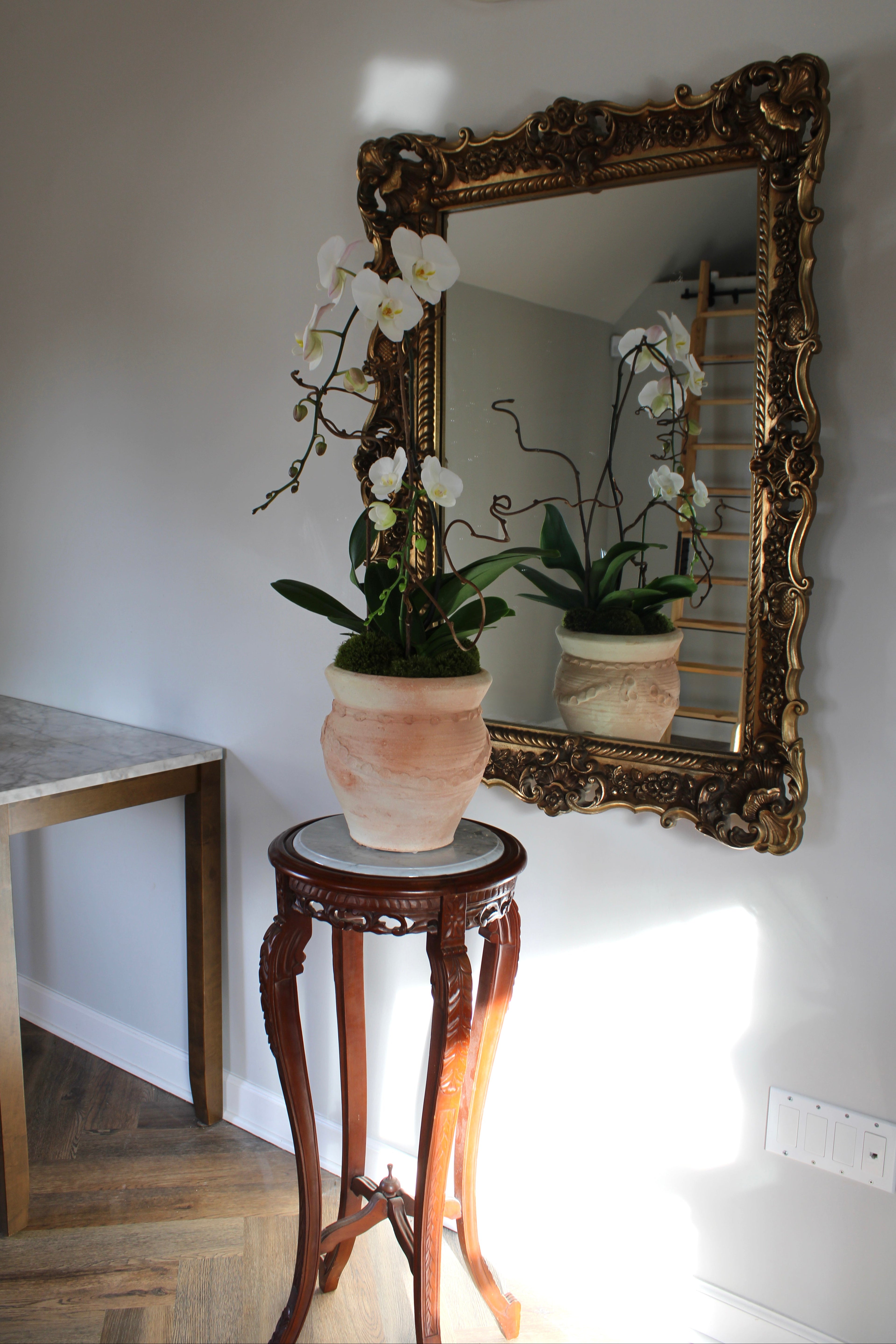 Decorative mirror with gold frame reflecting a plant on a small table against a white wall.